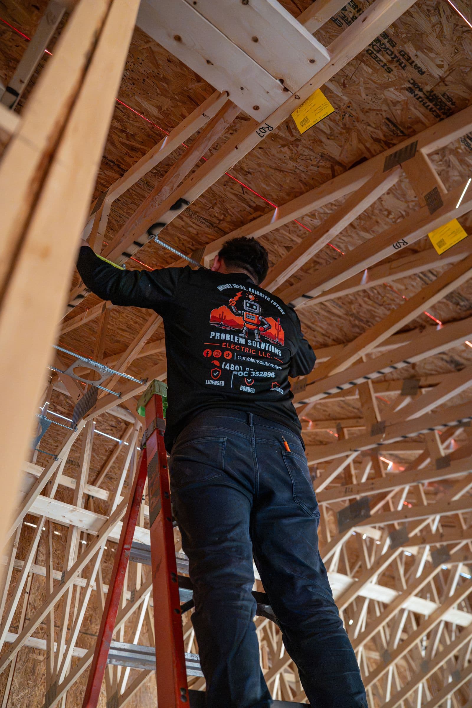 A technician on a ladder in the trusses, the robot logo on his shirt